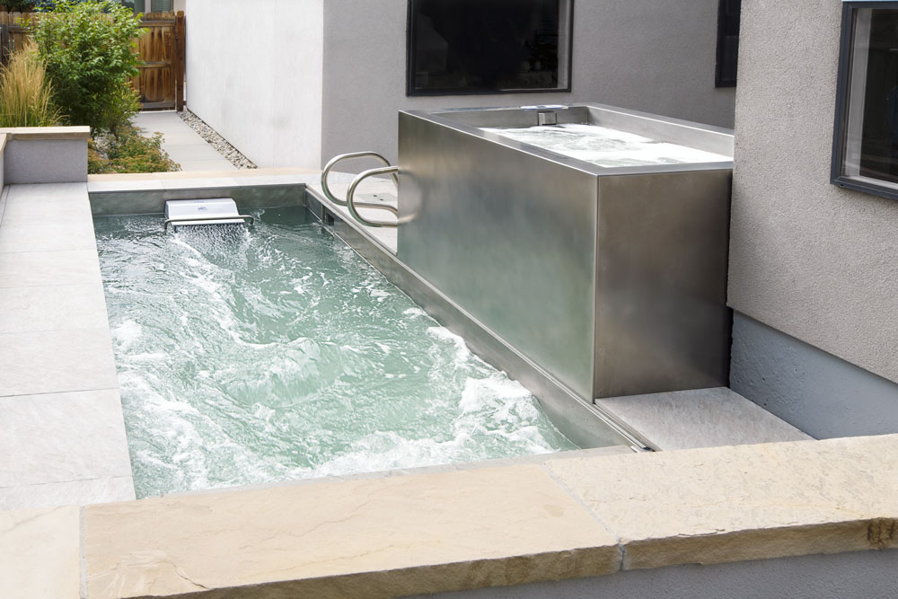 Stainless steel swim spa with LED lighting and automatic cover.  180”x84”x58”.  Located above is a stainless steel spa with bench seating and LED lighting.  48”x 70”x 34”