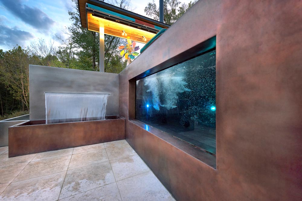 Custom copper spa with glass side window neighboring custom stainless steel spa with water feature and catch basin.