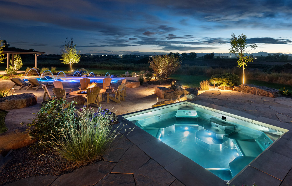 Stainless steel spa with four captain chairs, bench seating, cool down area, LED lighting and automatic cover. 115”x143”x 46”.In background is a stainless-steel pool with sun shelf, bench seating on side walls of pool, swim up bar, swim spa, laminar jets, and automatic cover.  193”x402”x69”